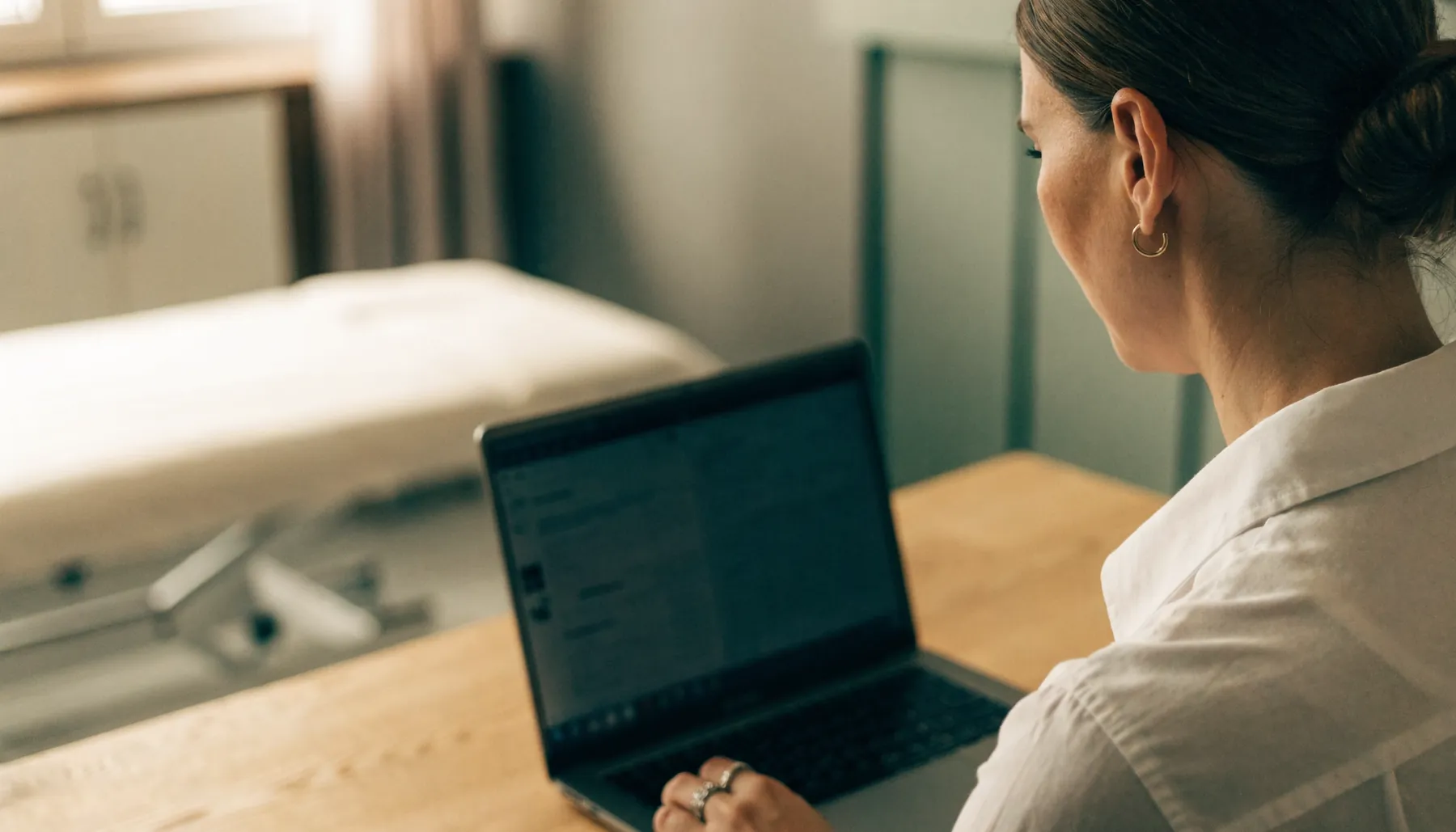 A physiotherapist reviewing a patient report on a laptop in a clinic treatment room.