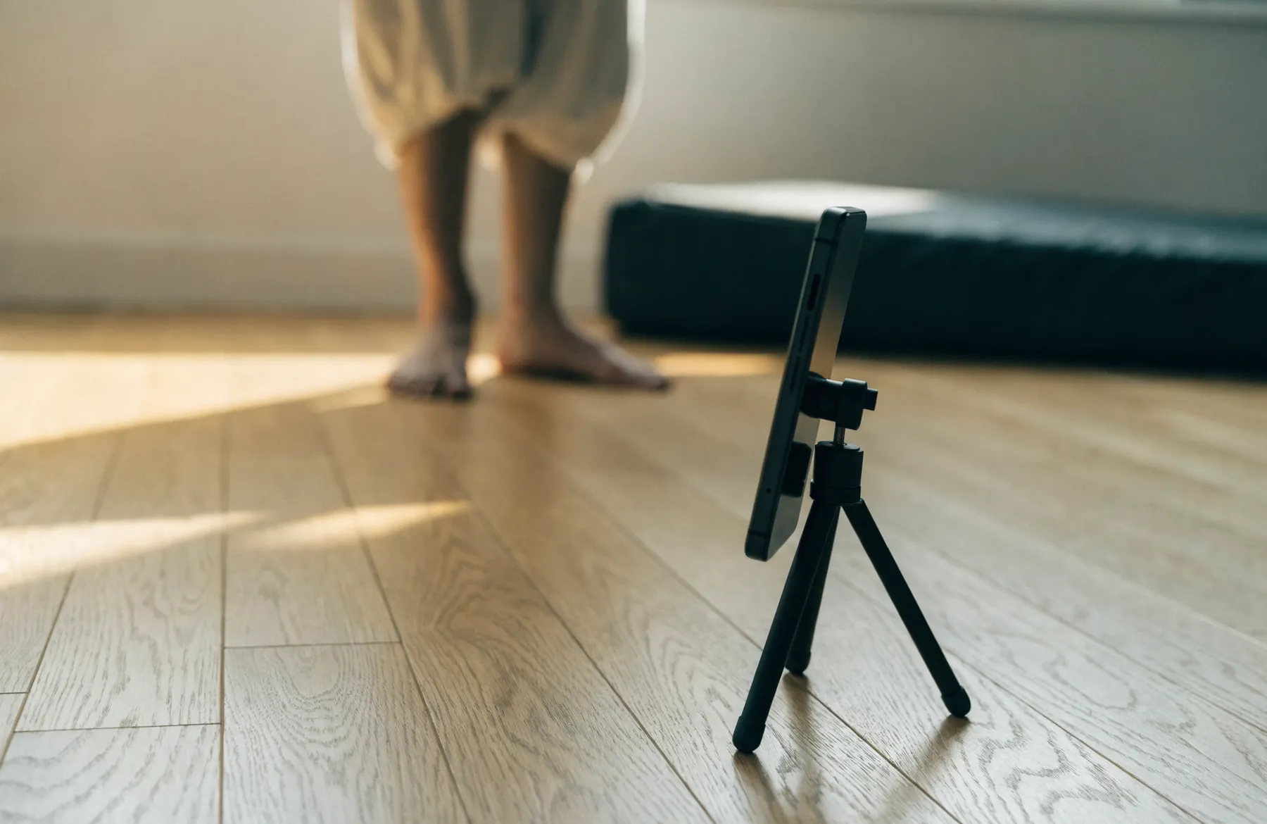 A phone standing in a small tripod on a wooden floor at floor level, with bare feet blurred in the background.