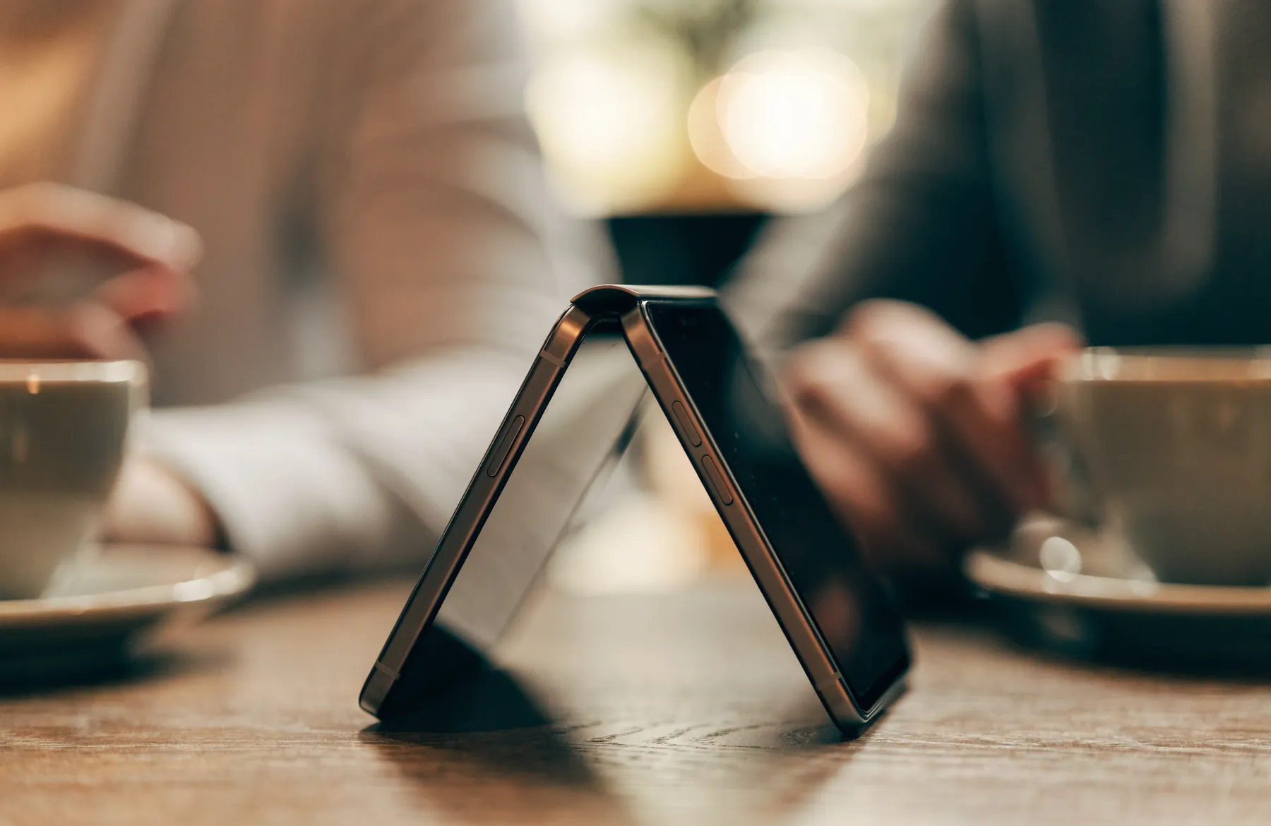 A foldable phone standing open in tent posture on a wooden cafe table between two seated people.