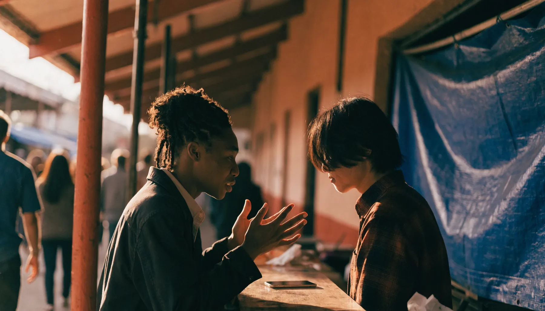 Two people talking across a counter at a covered market in warm afternoon light, with a phone lying flat on the counter between them.